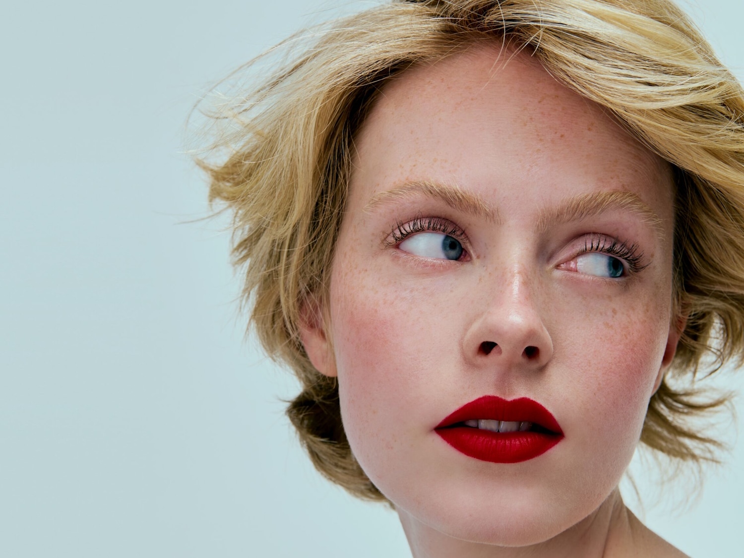 Nahaufnahme eines hellhäutigen Models mit blonden, gewellten Haaren, Sommersprossen und leuchtend rotem Lippenstift, das zur Seite blickt.