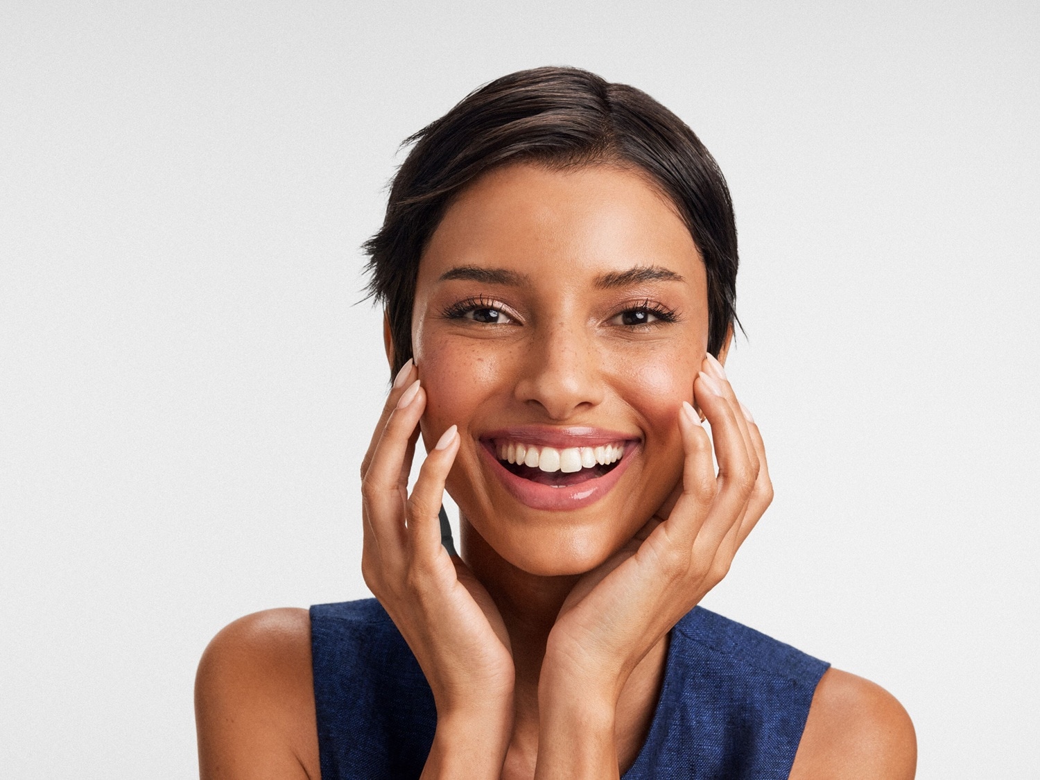 Eine strahlende Frau mit kurzem, dunklem Haar und einem blauen Oberteil lächelt und berührt mit beiden Händen ihr Gesicht.