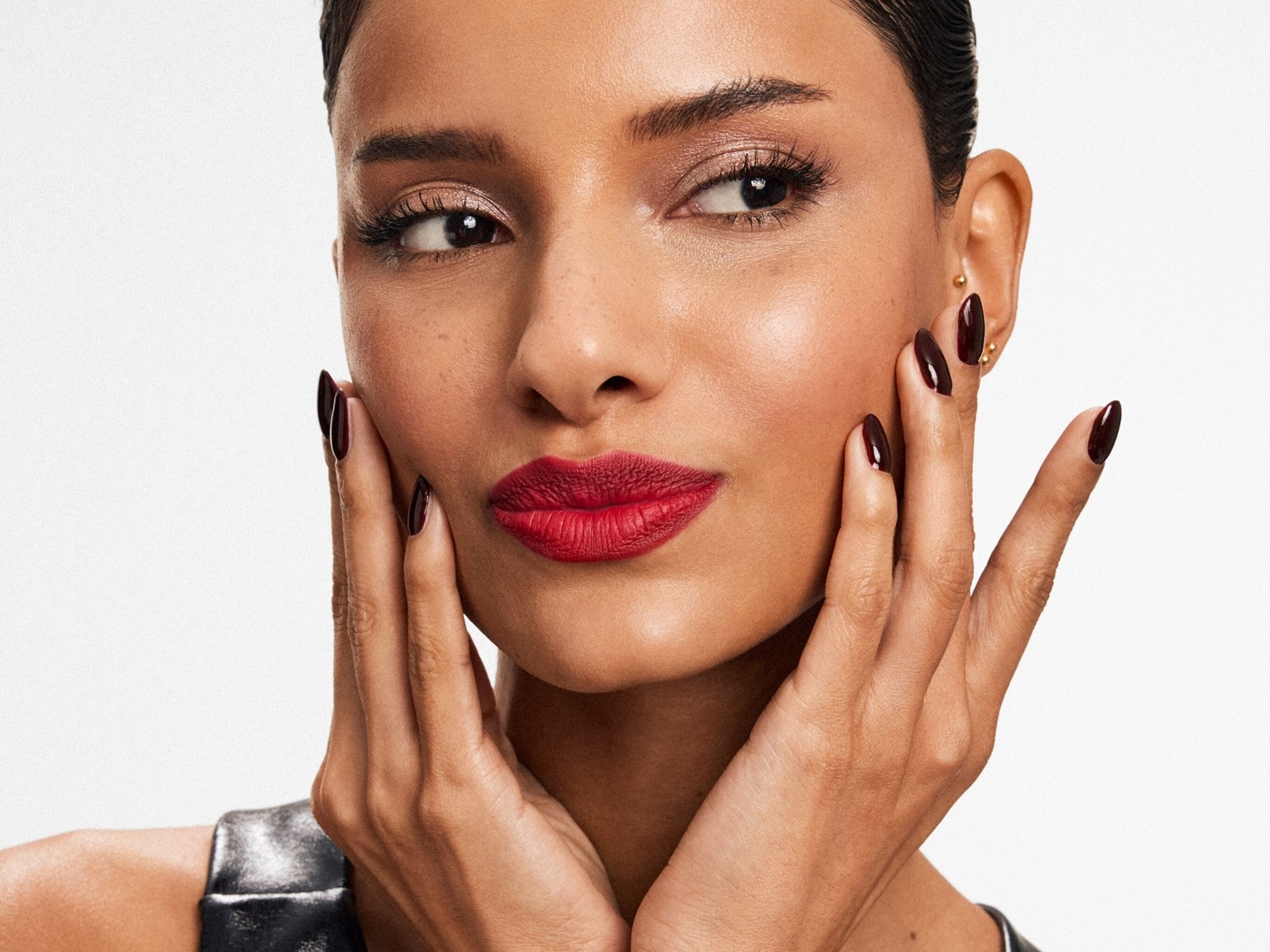 Nahaufnahme einer Frau mit roten Lippen und dunkelroten Nägeln, die ihr Gesicht berührt und einen glamourösen Look zeigt.