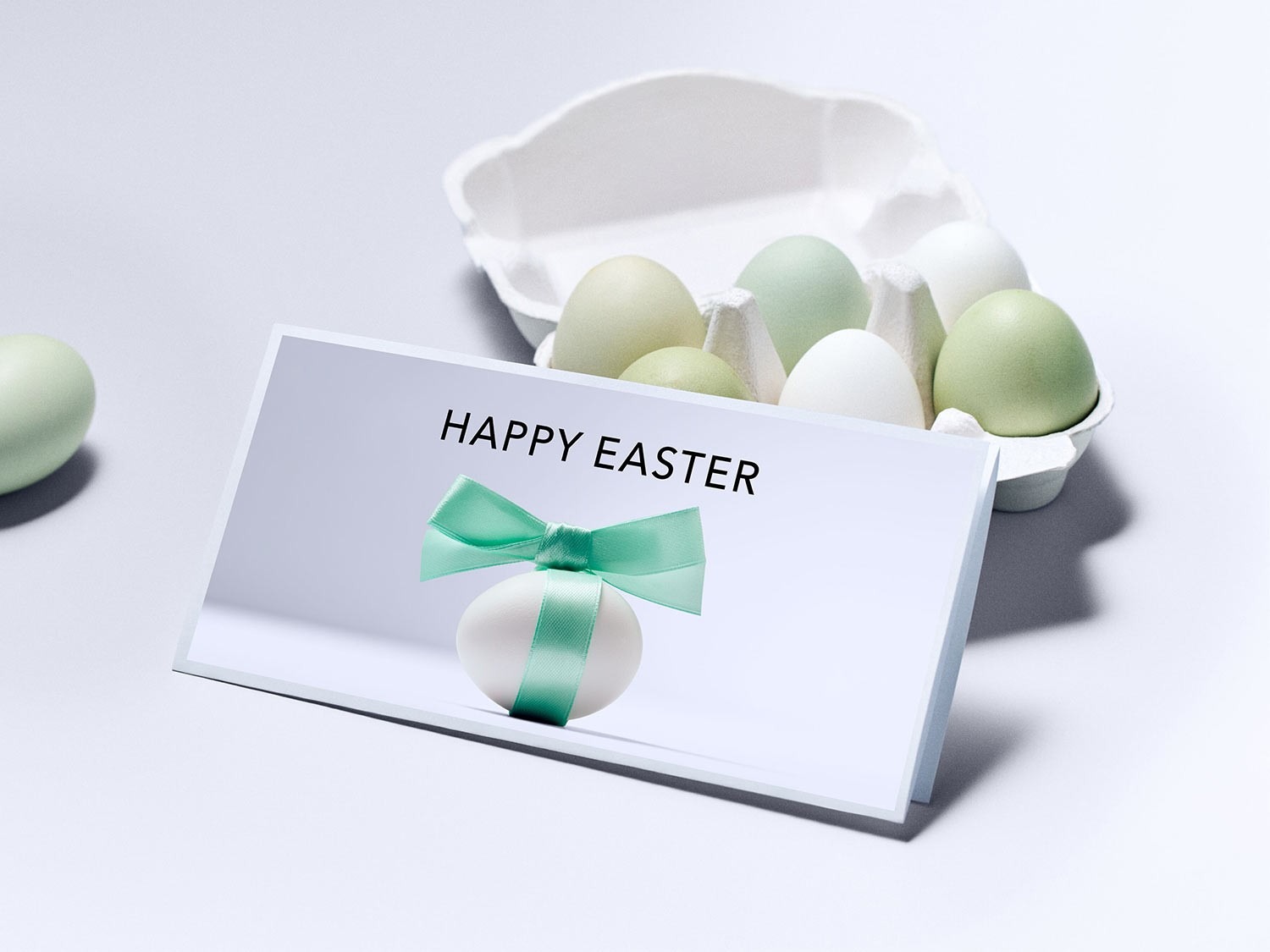 Eine weiße Karte mit der Aufschrift "Happy Easter" liegt vor einer Eierpackung mit grün und weiß gefärbten Eiern, die mit einer Schleife verziert ist.