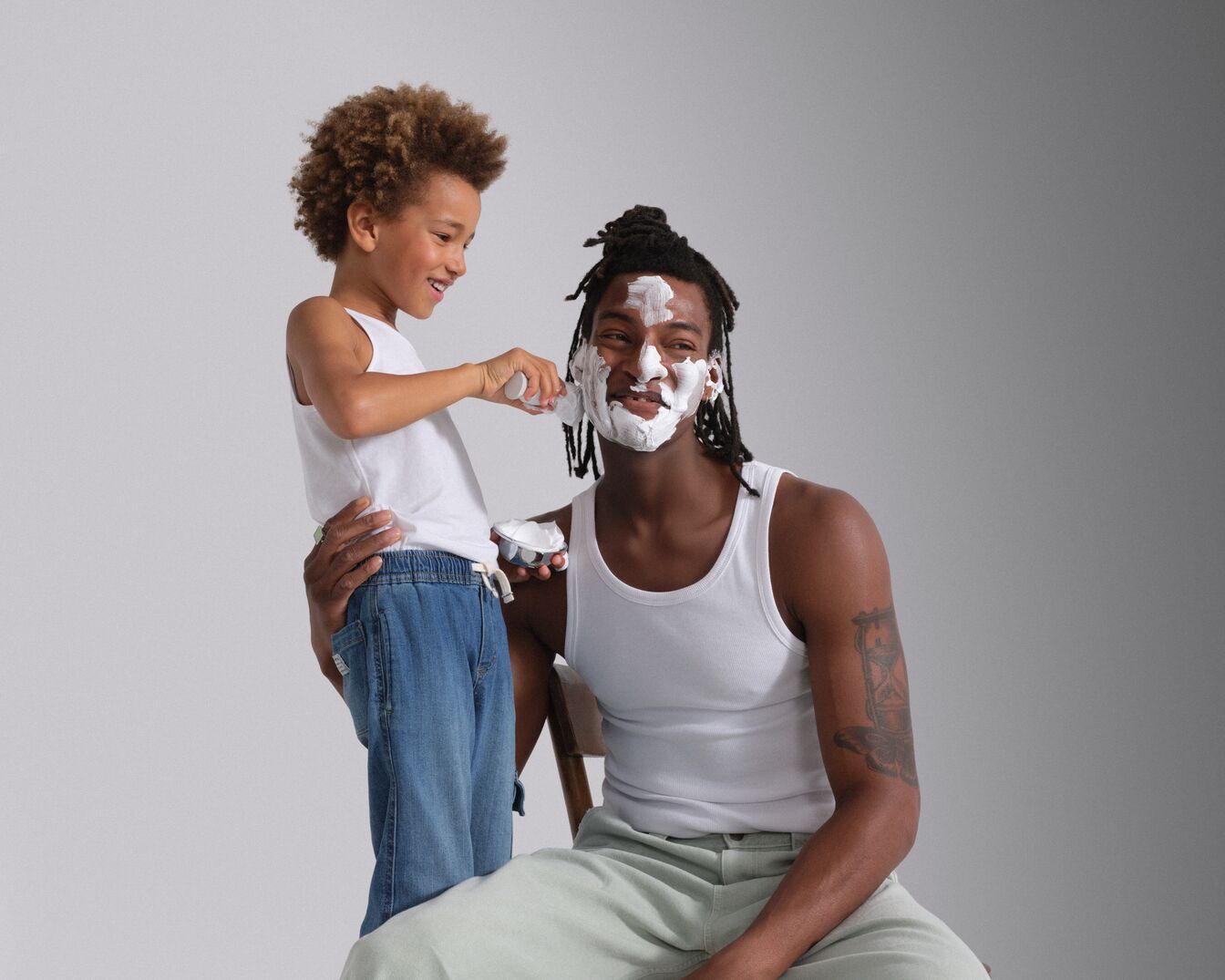 Un jeune garçon met de la crème à raser blanche sur le visage de son père, tous deux souriant et portant des débardeurs blancs.