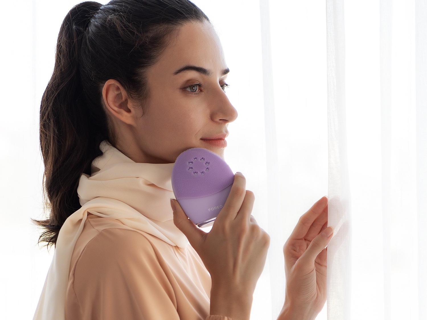 Une femme tient un appareil de nettoyage pour le visage violet de marque Foreo, se préparant à l'utiliser pour une peau éclatante.