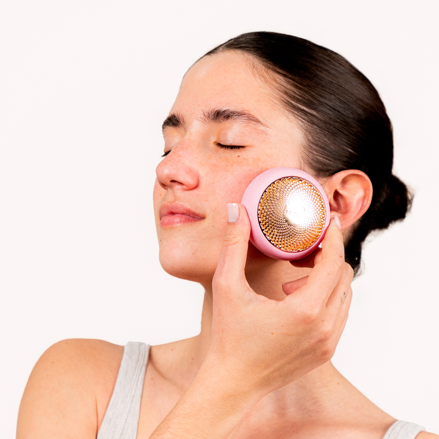 Une femme utilise un appareil de soin de la peau rose et doré sur sa joue, les yeux fermés, pour un moment de détente et de beauté.