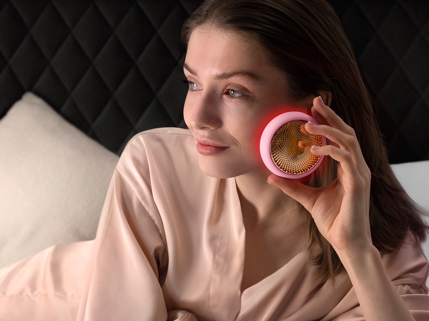 Une femme en chemise de nuit pêche utilise un appareil de beauté rose et or sur sa joue, illuminant sa peau d'une douce lueur.