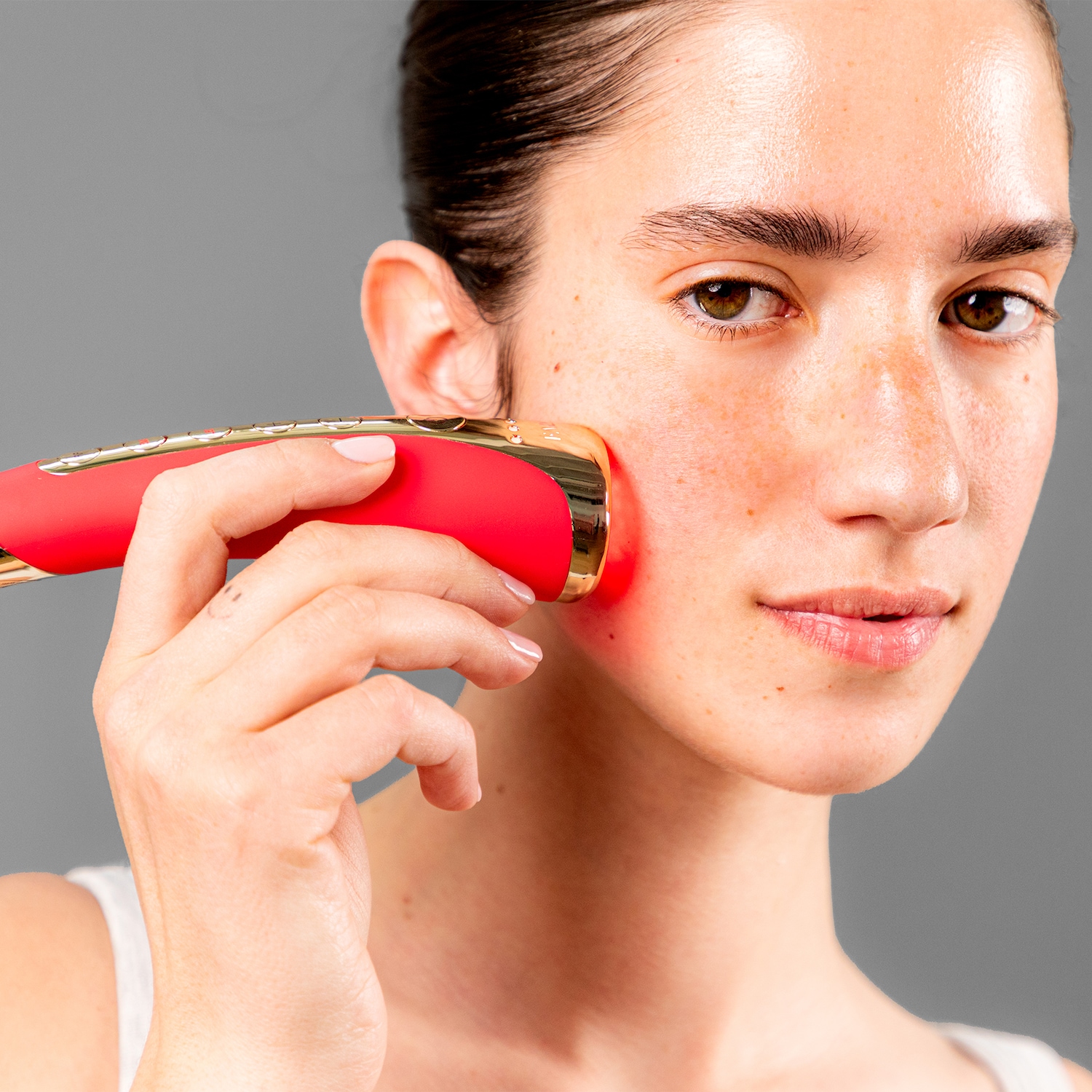 Une femme utilise un appareil de beauté rouge et doré sur sa joue pour un soin de la peau ciblé.