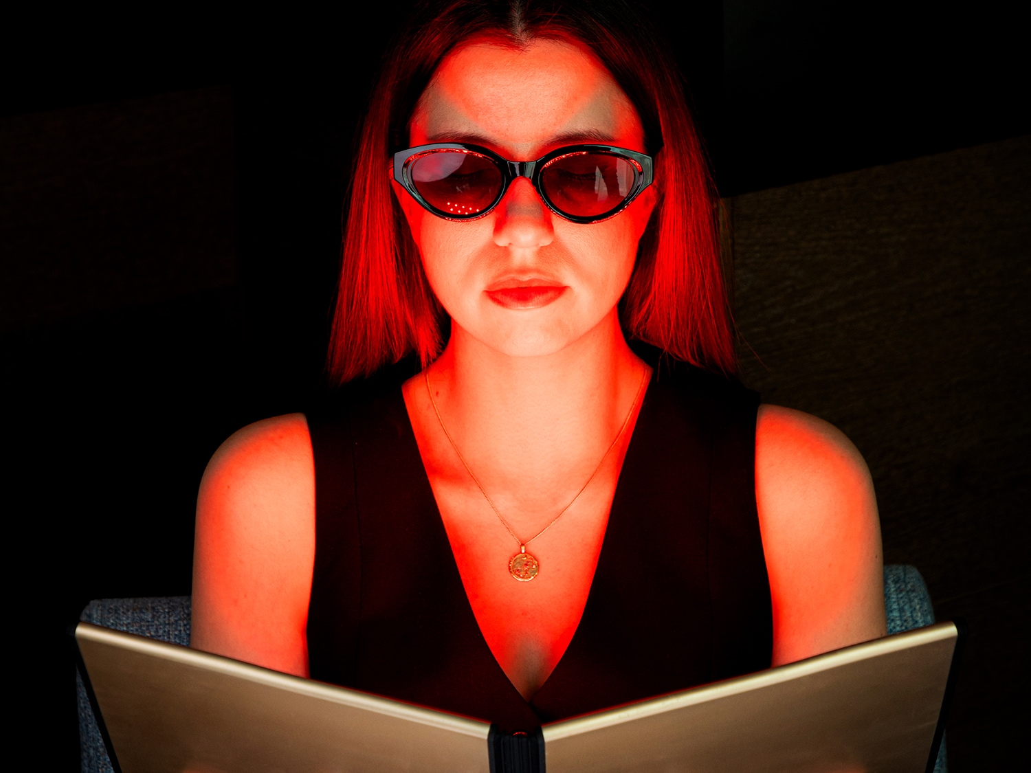 Une femme portant des lunettes noires et un haut noir regarde un livre dans une lumière rouge.