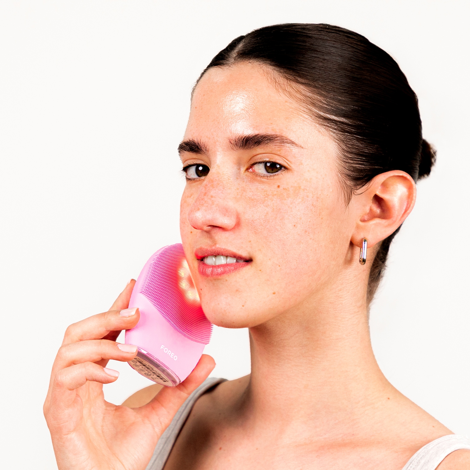 Une femme utilise un appareil de nettoyage pour le visage rose de la marque FOREO, qui émet une lumière rouge douce sur sa peau, pour un teint éclatant et soigné.
