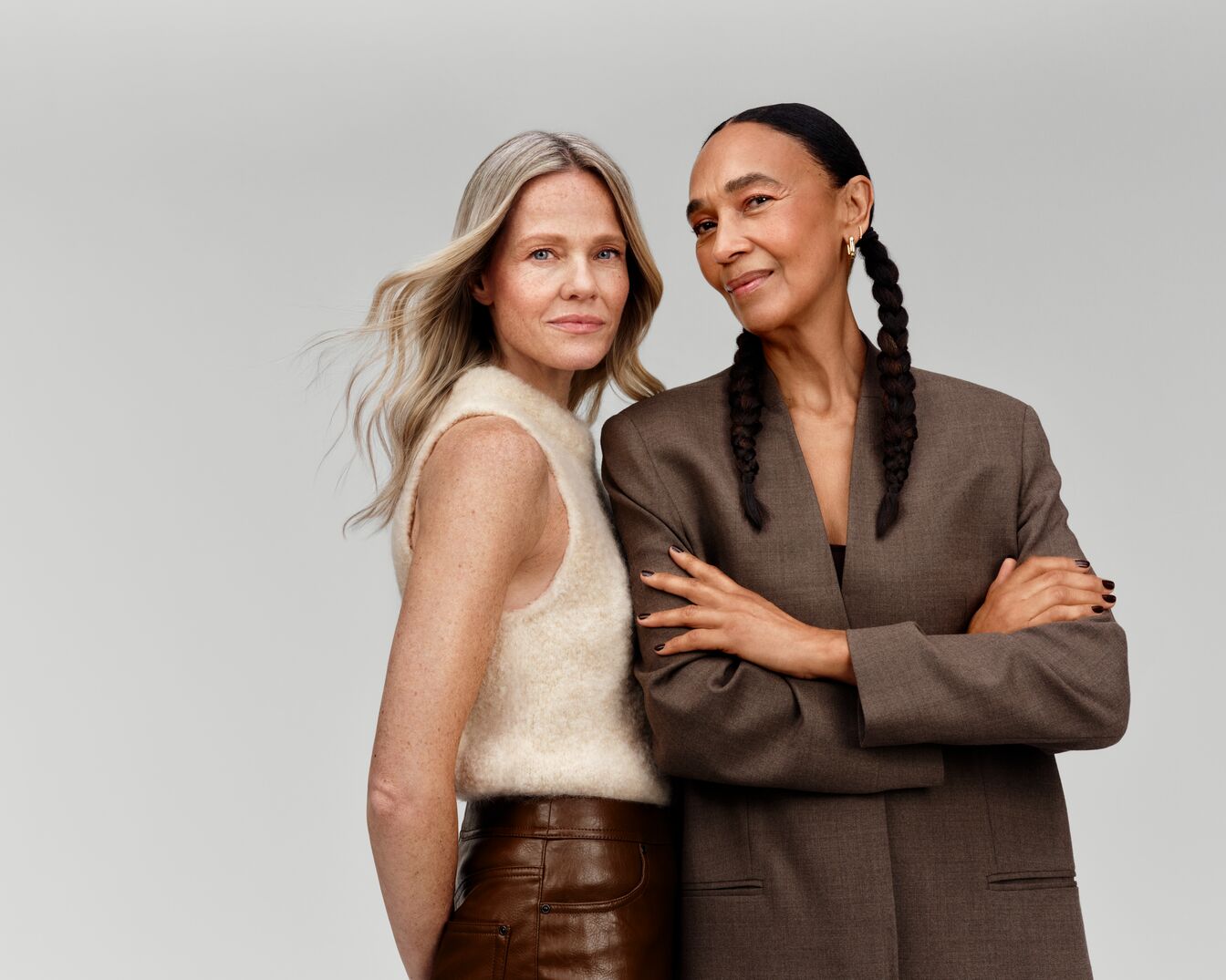 Zwei Frauen posieren selbstbewusst: Die blonde Frau trägt einen cremefarbenen Pullunder und eine braune Lederhose, während die dunkelhäutige Frau in einem braunen Blazer mit geflochtenen Zöpfen zu sehen ist.
