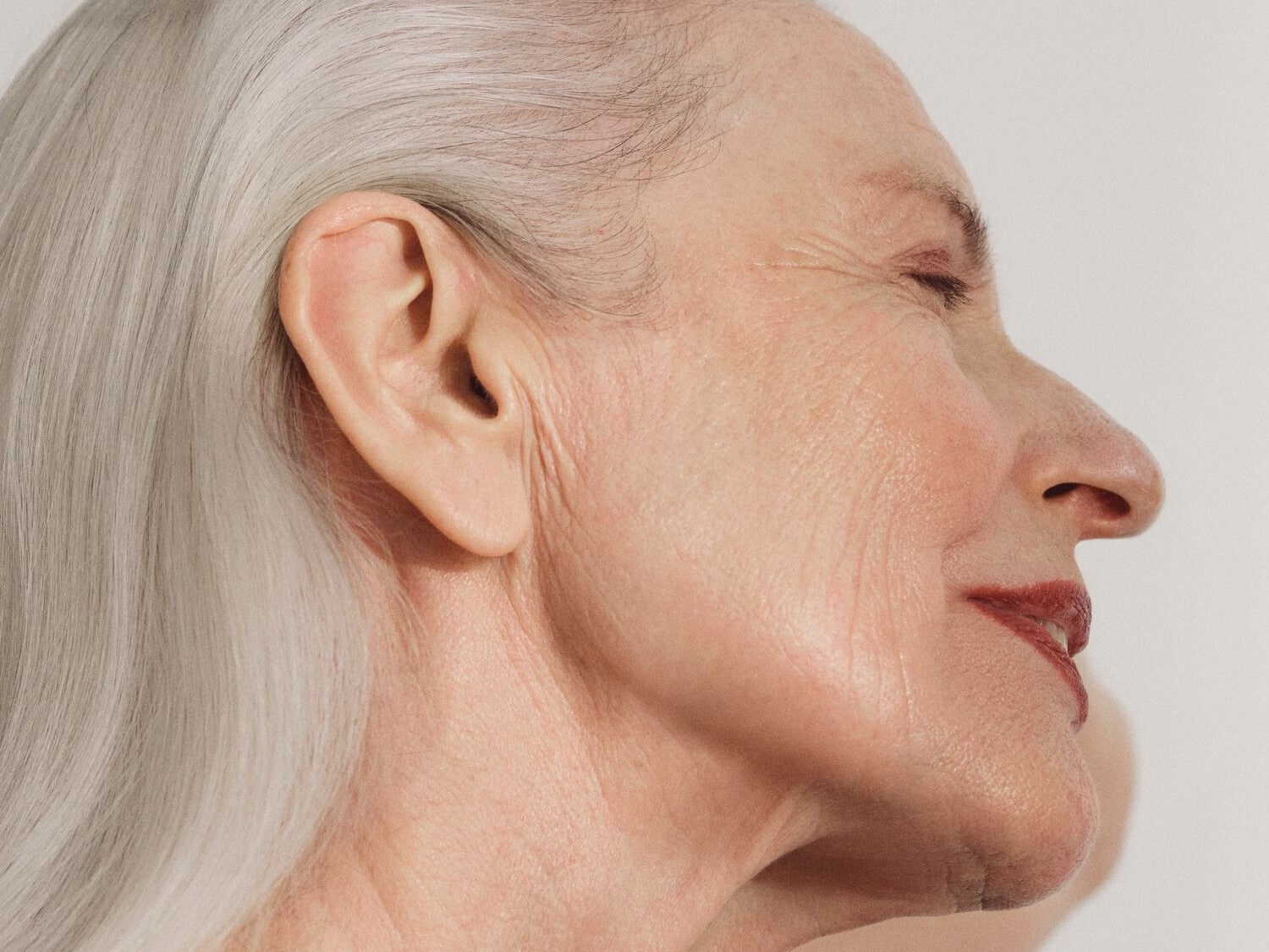 Profil einer älteren Frau mit grauen Haaren und roten Lippen, die ihre Augen geschlossen hat, was die natürliche Schönheit und Würde des Alterns hervorhebt.