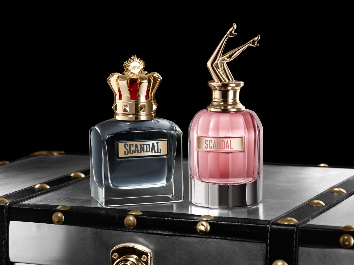 Dva flakóny vôní Scandal, jeden s korunou a tmavomodrým odtieňom, druhý s nohami a ružovým odtieňom, s nápisom Scandal na oboch, sedia na striebornom kufri.