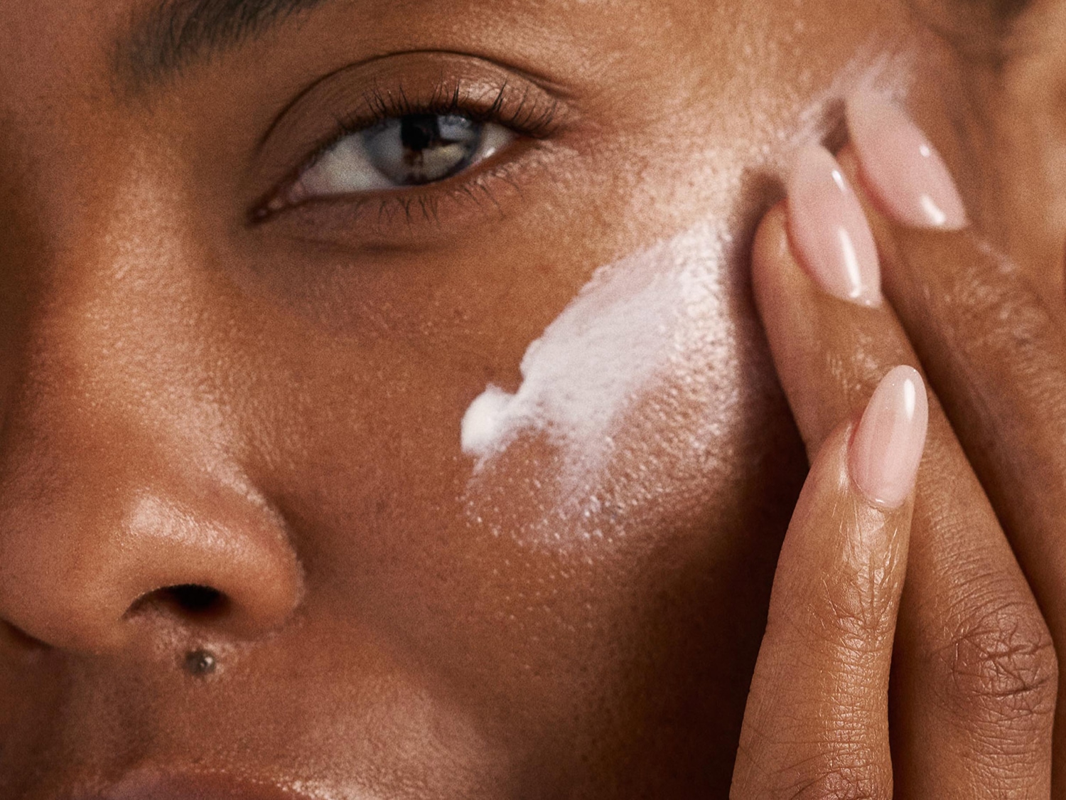 Close-up van een vrouw met een getinte huid die een witte crème aanbrengt op haar gezicht met haar vingers, haar nagels zijn lichtroze gelakt.