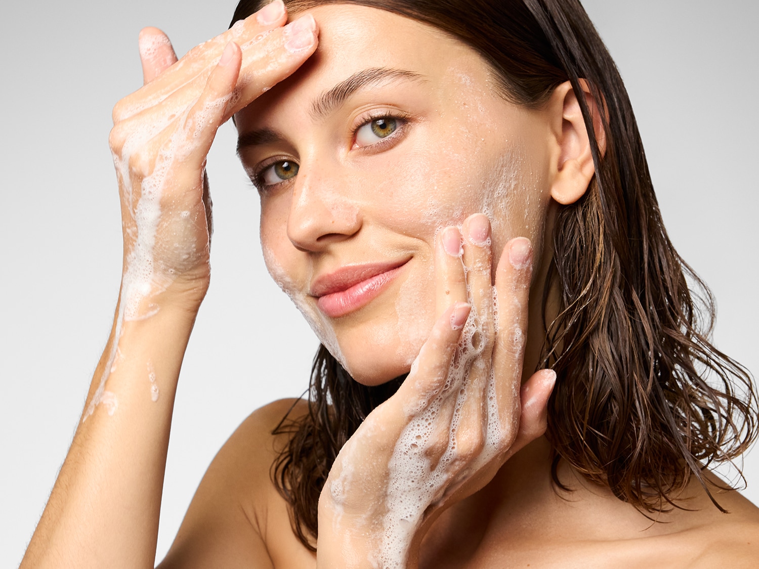 Een vrouw met nat haar masseert schuimend reinigingsmiddel op haar gezicht met een glimlach.