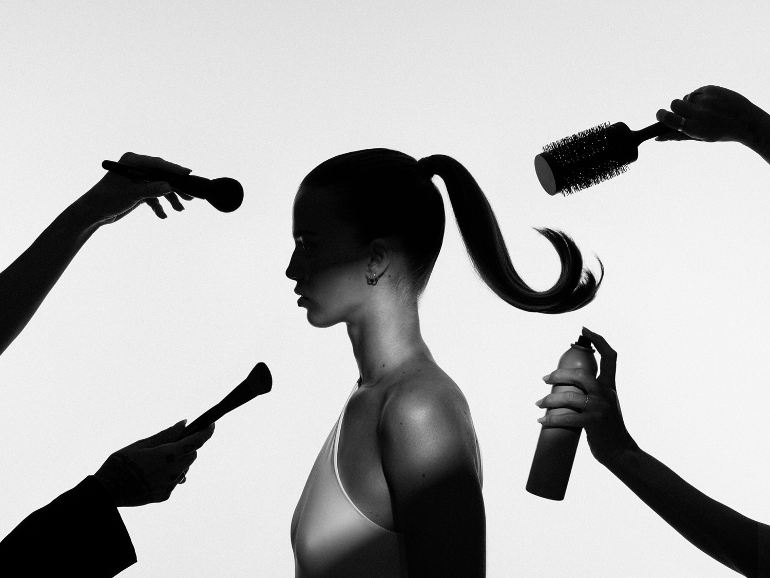 Une femme de profil se fait coiffer et maquiller par plusieurs mains tenant des pinceaux, une brosse ronde et une laque, le tout en noir et blanc.