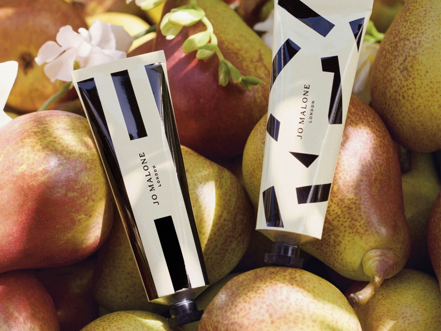 Deux tubes de crème pour les mains Jo Malone London, ornés de motifs géométriques noirs, reposent au milieu de poires mûres et de fleurs blanches.