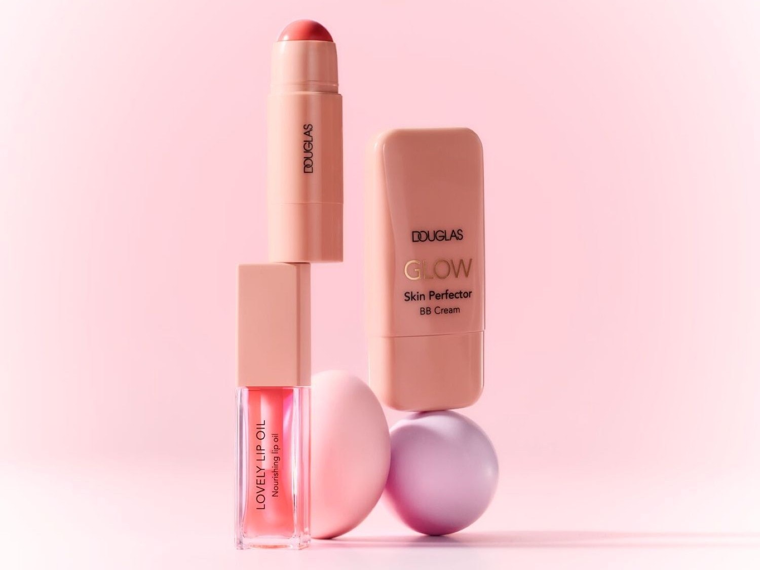 Su sfondo rosa chiaro, una composizione di prodotti Douglas color pesca: un olio labbra Lovely Lip Oil, uno stick labbra e un BB cream Glow Skin Perfector.