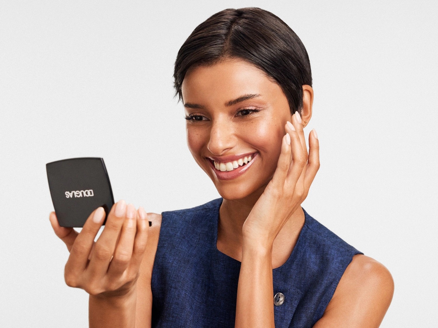 Une femme souriante, vêtue d'une robe bleue, tient un poudrier noir de marque Douglas et touche son visage radieux.