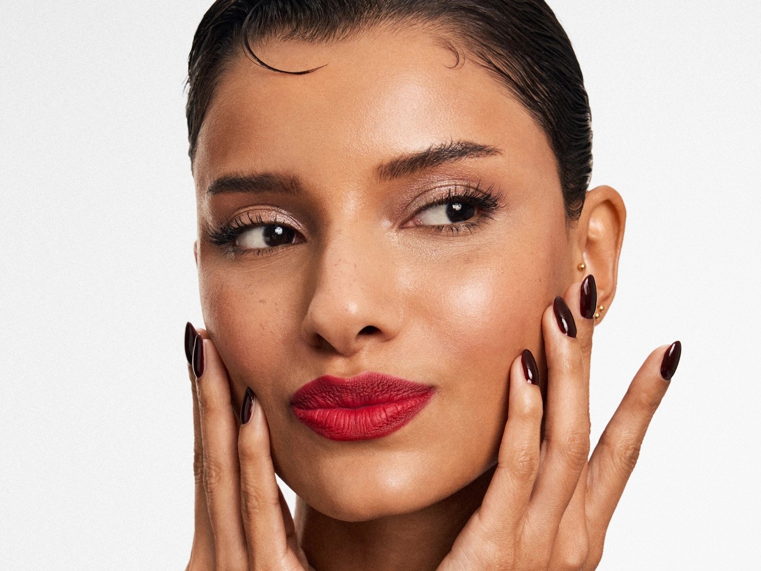 Gros plan sur un visage féminin avec un rouge à lèvres rouge vif et des ongles bordeaux brillants, les mains touchant délicatement le visage, mettant en valeur un maquillage élégant et une manucure soignée.