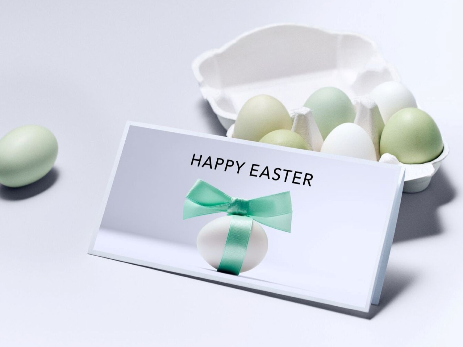 Una confezione di uova bianche e verdi pastello è esposta accanto a un biglietto d'auguri con la scritta 'Buona Pasqua' e decorato con un uovo avvolto in un nastro verde.