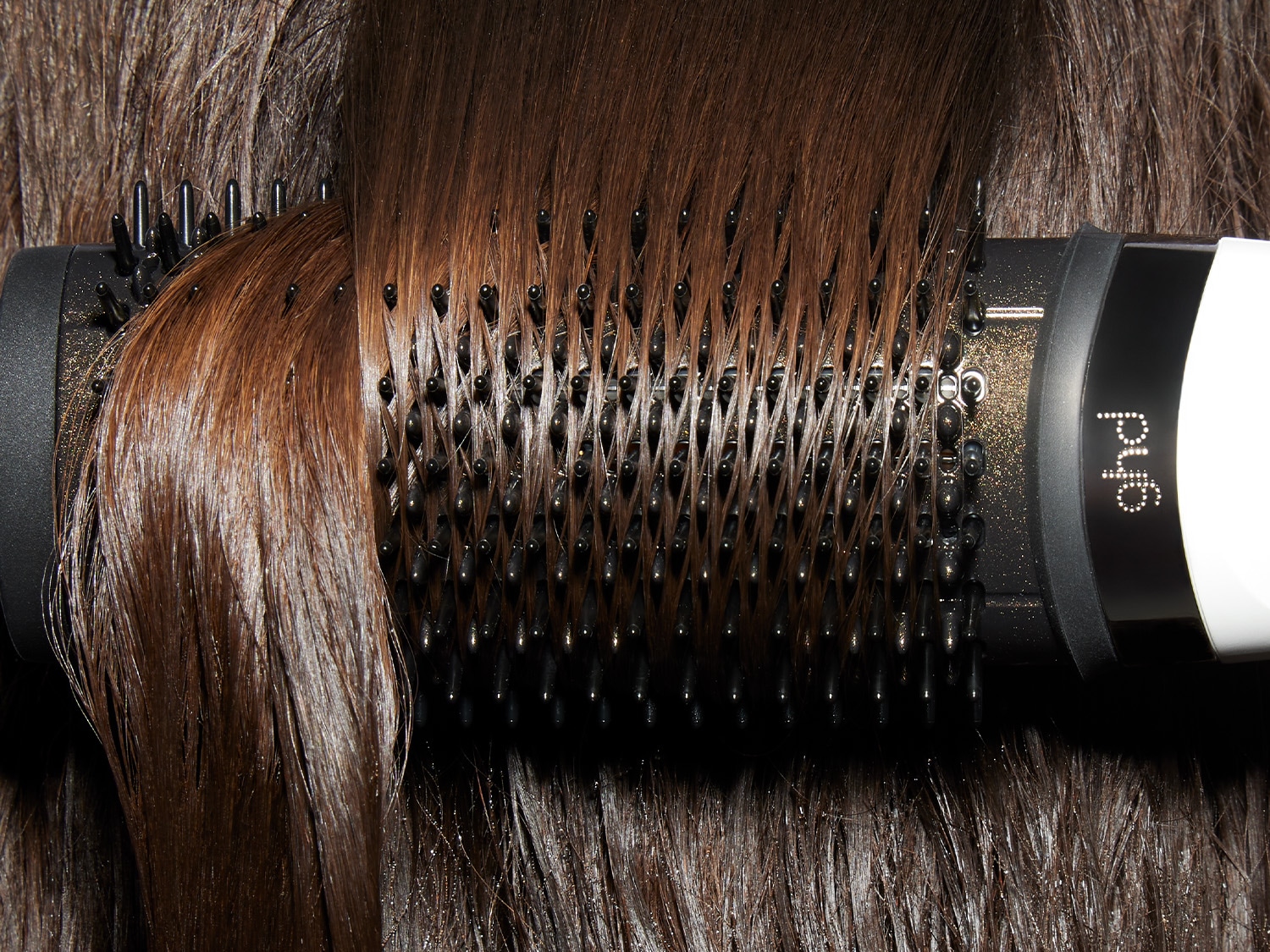 Nahaufnahme einer braunen Haarsträhne, die um eine schwarze Rundbürste mit dem Schriftzug ghd gewickelt ist, um das Haar zu stylen.