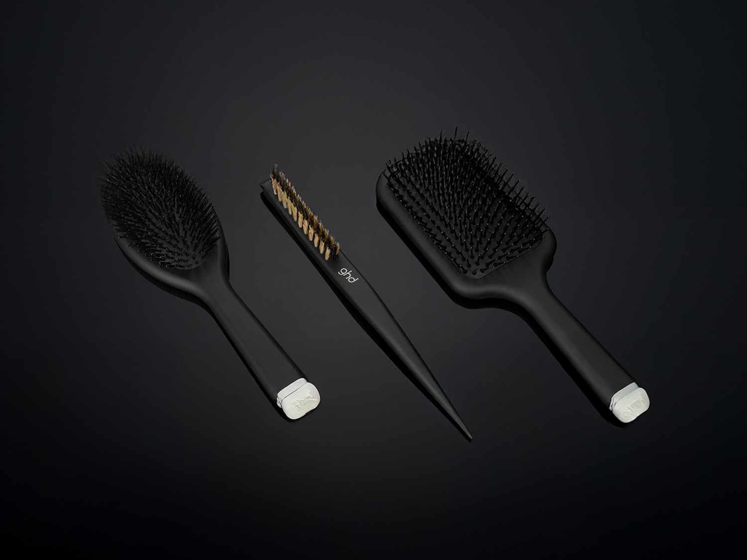 Drei schwarze ghd Haarbürsten, darunter eine Paddle Brush, eine Oval Dressing Brush und ein Stylingkamm, liegen auf einem glänzenden schwarzen Untergrund.