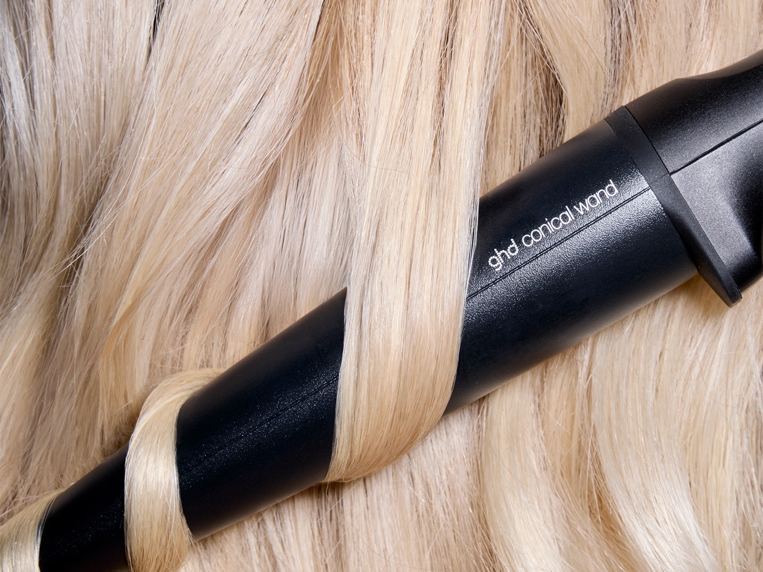 Nahaufnahme eines schwarzen ghd konischen Zauberstabs, der in blondes Haar eingewickelt ist und Locken erzeugt.