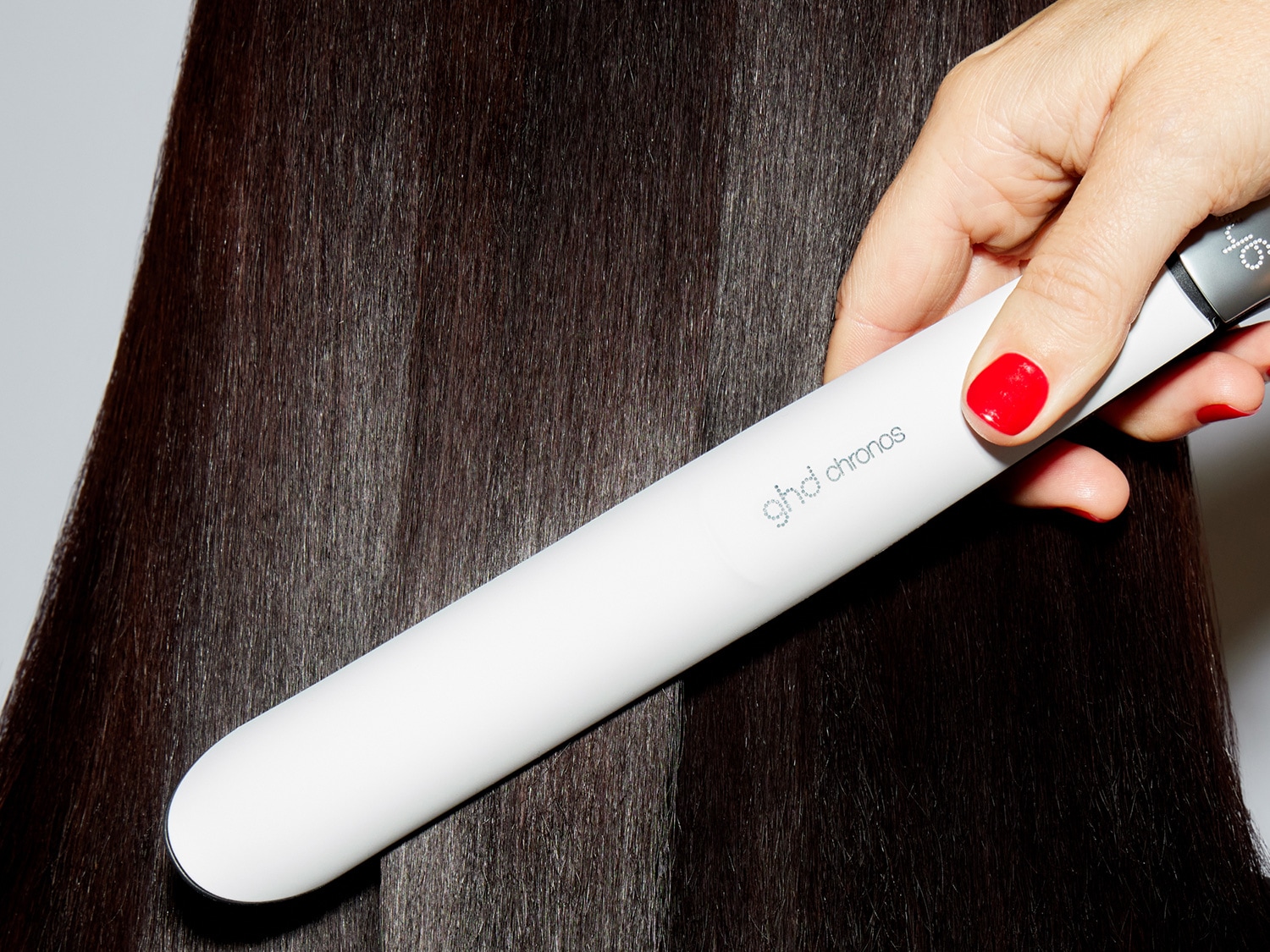 Eine Hand mit rot lackierten Nägeln hält ein weißes ghd Chronos Glätteisen, das durch dunkles, glattes Haar gleitet.
