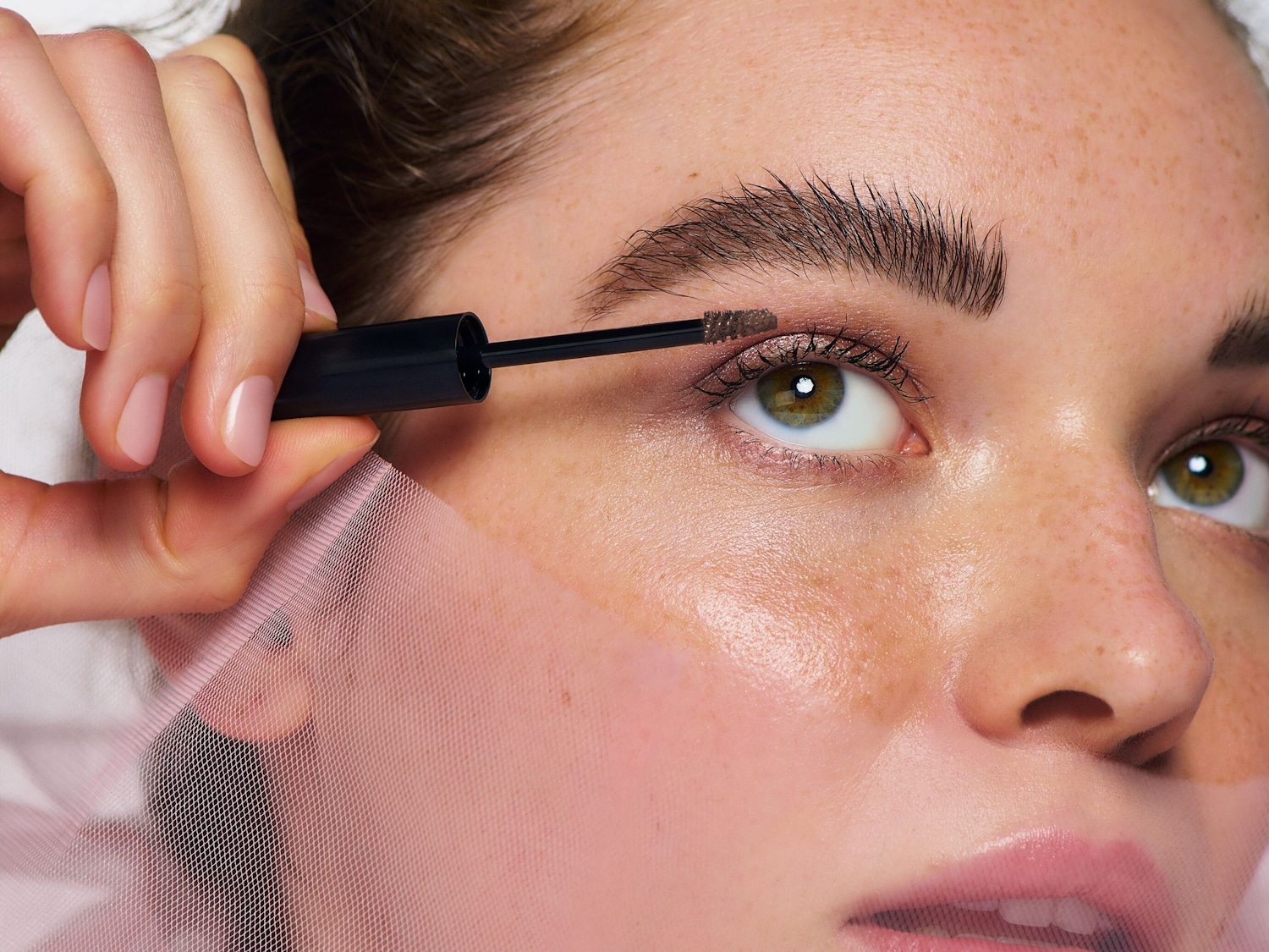 Una donna con lentiggini e occhi verdi applica un mascara marrone sulle sopracciglia folte con un applicatore nero, mentre una rete rosa copre parte del suo viso.