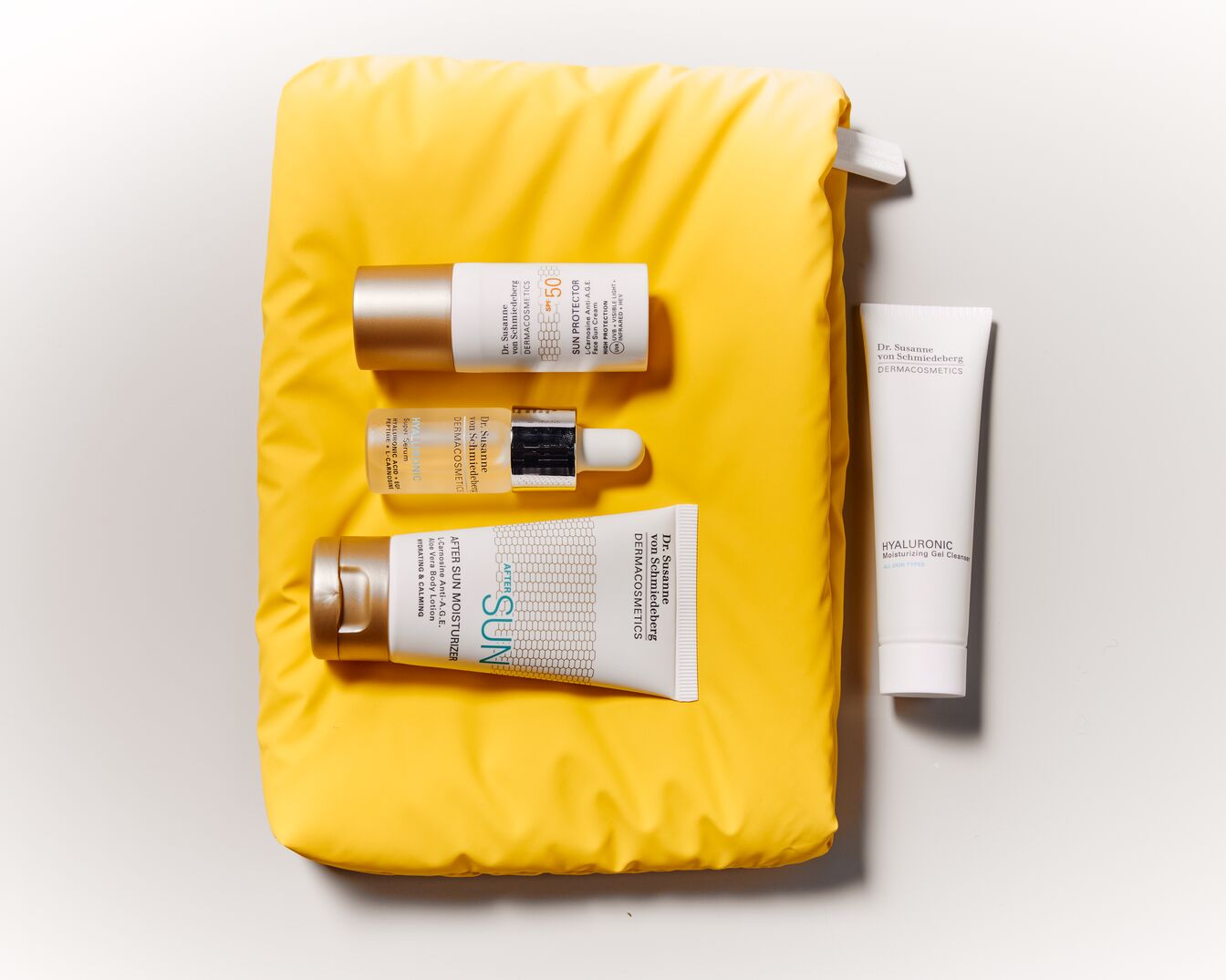 Žlutá kosmetická taška s opalovacím krémem Dr. Susanne von Schmiedeberg DERMACOSMETICS s SPF 50, sérem s kyselinou hyaluronovou, hydratačním přípravkem po opalování a hydratačním gelovým čisticím prostředkem.