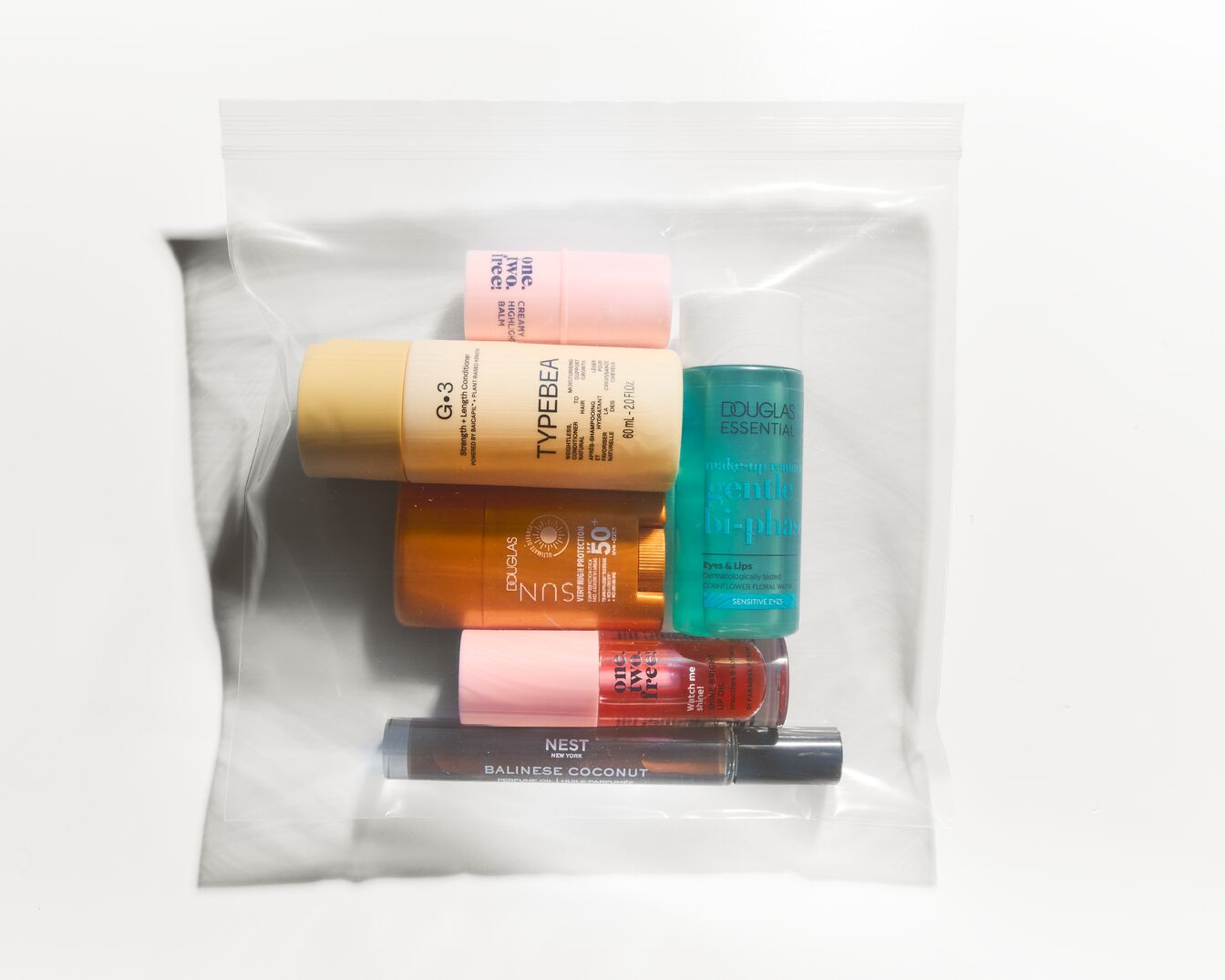 Průhledný plastový sáček obsahuje různé kosmetické produkty, včetně balzámu na rty, opalovacího krému Douglas SUN s SPF 50+, kondicionéru TYPEBEA a odličovače očí a rtů Douglas Essential.
