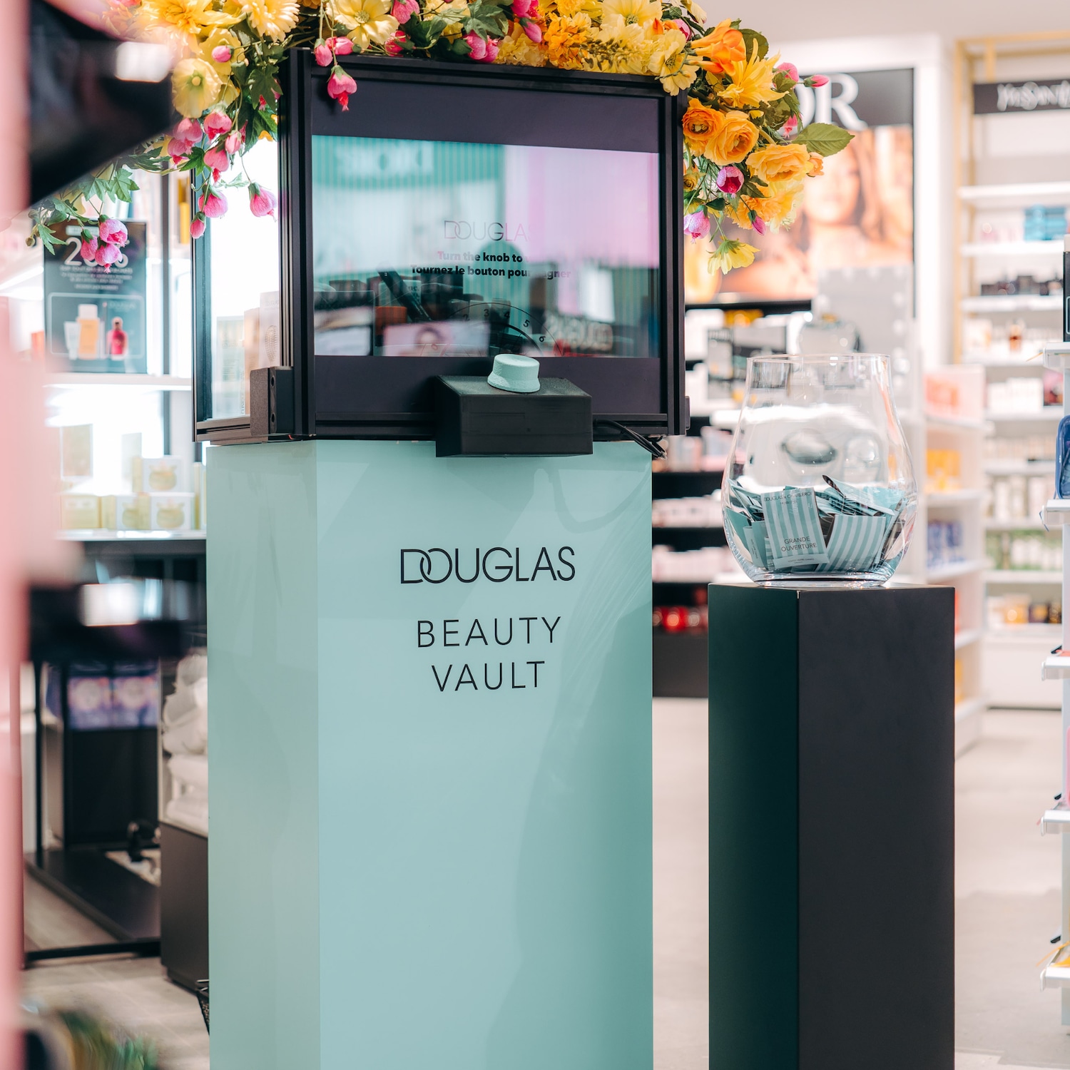 Découvrez le Beauty Vault de Douglas, orné de fleurs jaunes et roses, avec un écran interactif invitant à tourner le bouton pour gagner.