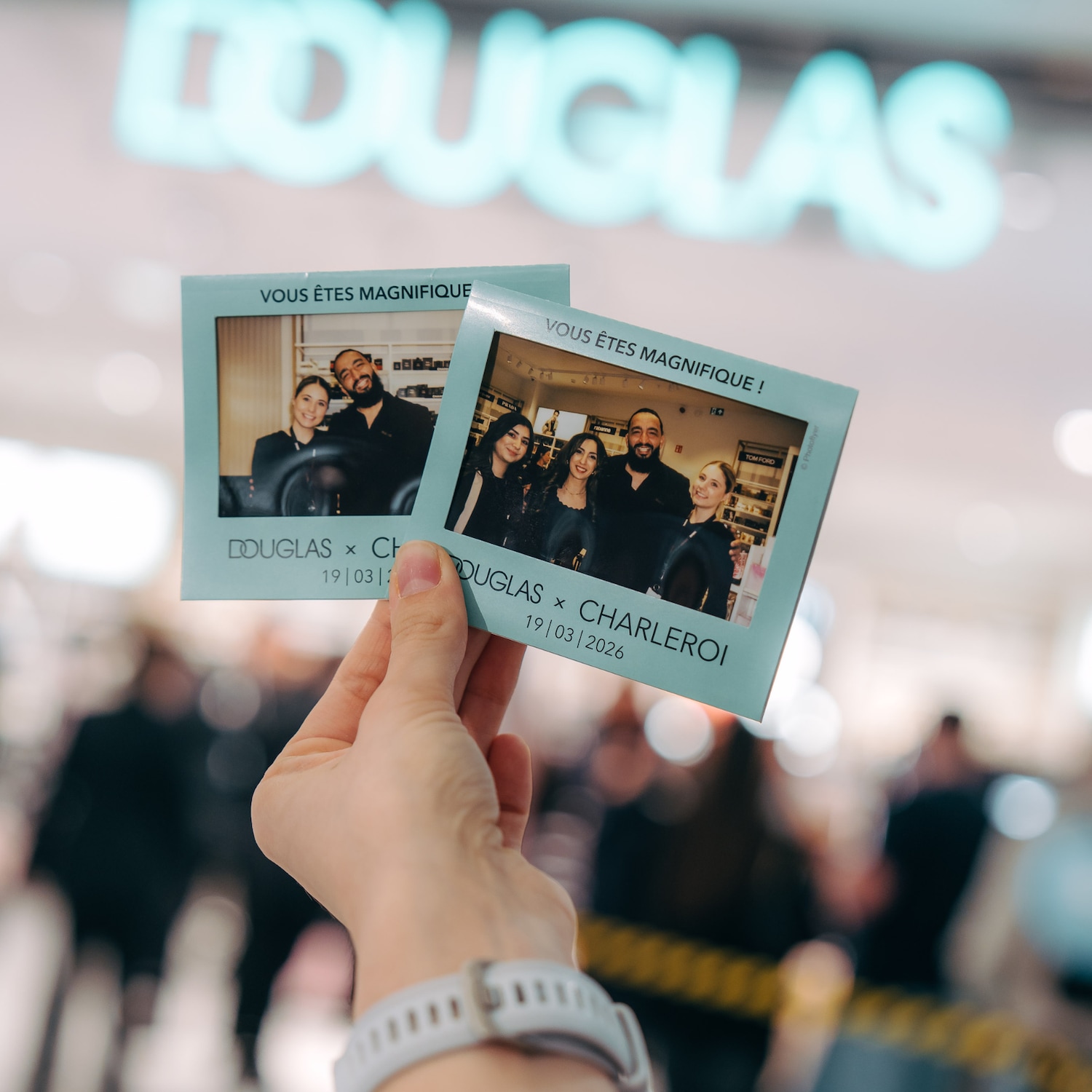 Une main tient deux photos de style polaroïd avec le texte 'VOUS ÊTES MAGNIFIQUE !' et 'DOUGLAS x CHARLEROI 19/03/2026', devant un logo Douglas flou.