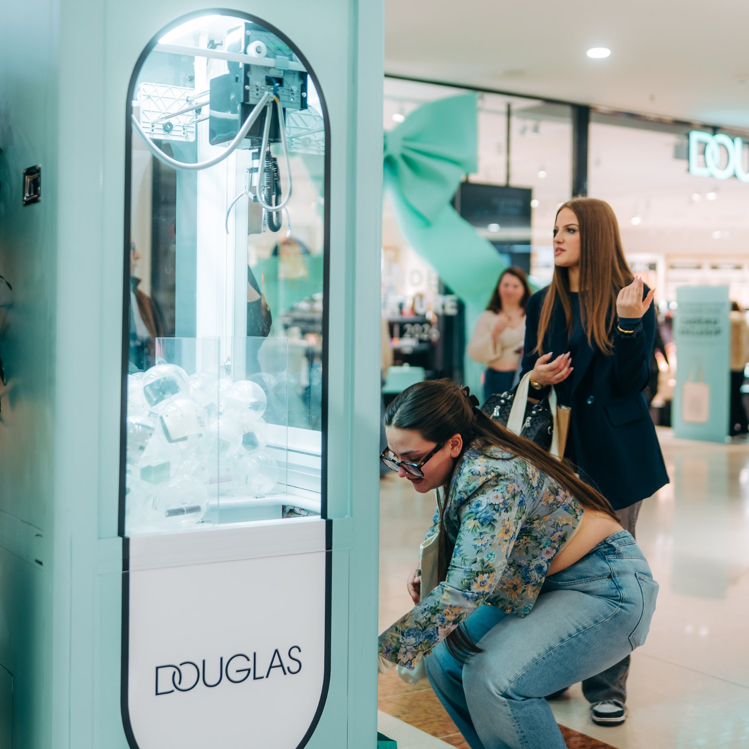 Une femme accroupie joue à une machine à pince de marque Douglas remplie de boules transparentes, tandis que deux autres femmes regardent.