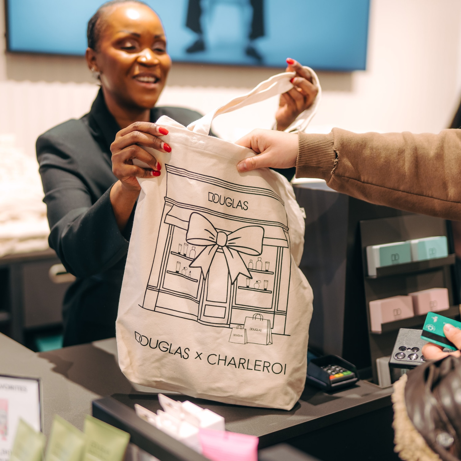 Une vendeuse souriante remet un sac en toile beige avec un dessin noir de la boutique Douglas et l'inscription « DOUGLAS x CHARLEROI » à un client payant avec une carte bancaire.