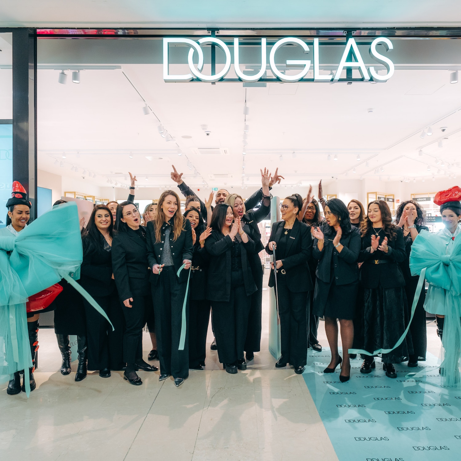 Devant le magasin Douglas illuminé, une équipe coupe un ruban bleu clair, célébrant l'ouverture avec des applaudissements et des sourires.