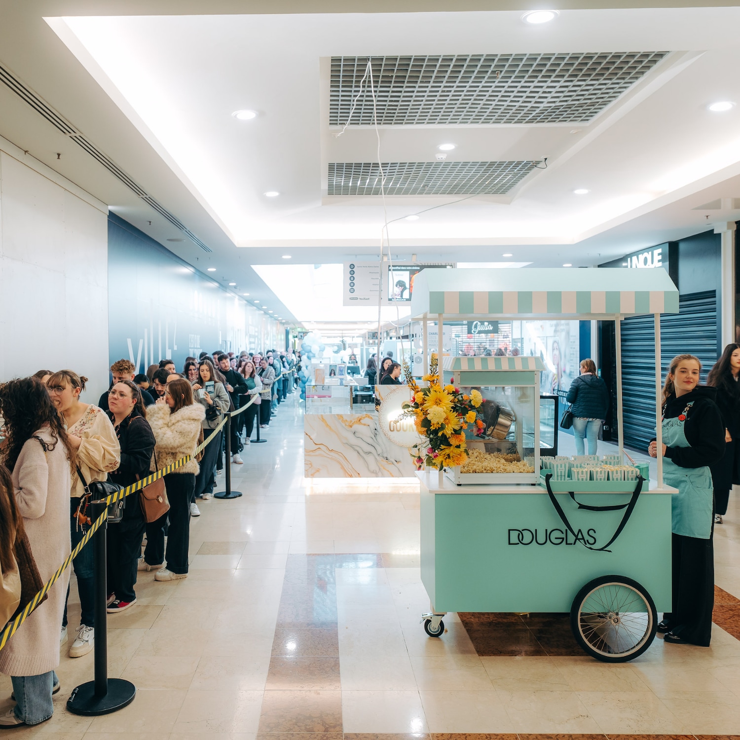 Une longue file de personnes attend devant un chariot bleu turquoise de Douglas, décoré de fleurs jaunes, dans un centre commercial lumineux.