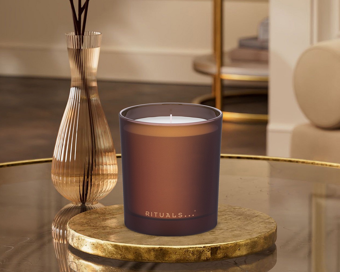 Ein braunes Rituals... Duftkerze sitzt auf einem goldenen Untersetzer, neben einer Vase mit Duftstäbchen, die eine warme und entspannende Atmosphäre schafft.