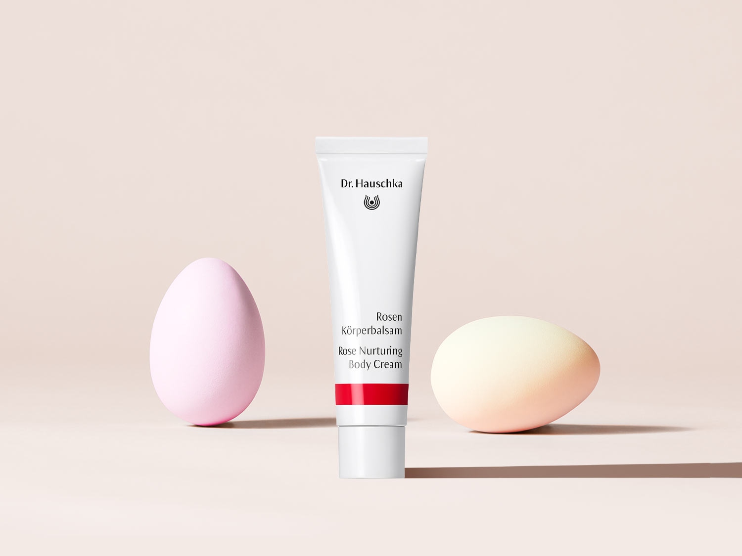 Dr. Hauschka Rosen Körperbalsam in einer weißen Tube mit roter Akzentfarbe, flankiert von zwei pastellfarbenen Ostereiern.