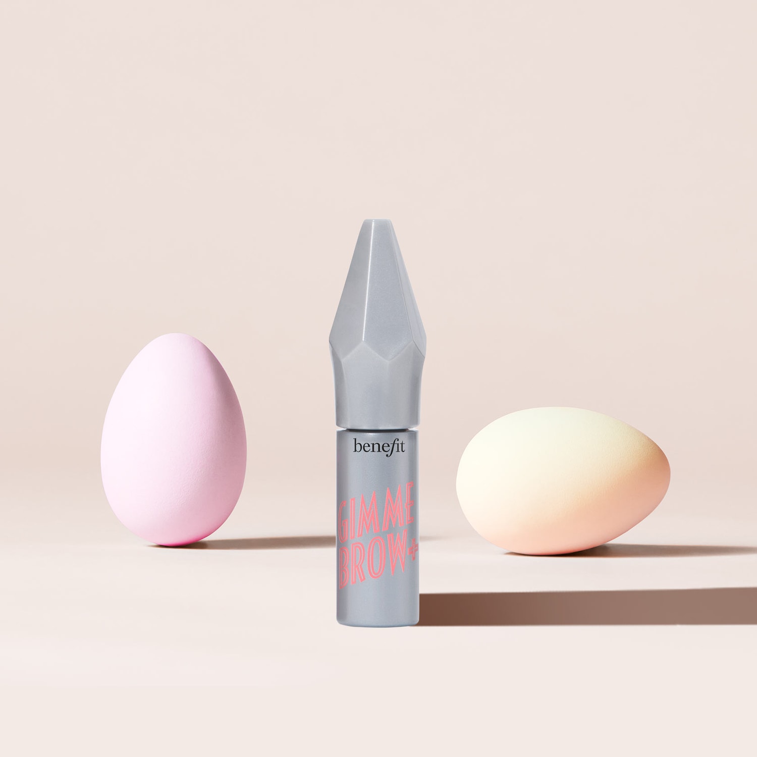Benefit Gimme Brow+ Volumen-Brauengel in grauer Verpackung mit rosa Schrift, flankiert von einem rosa und einem gelben Ei.