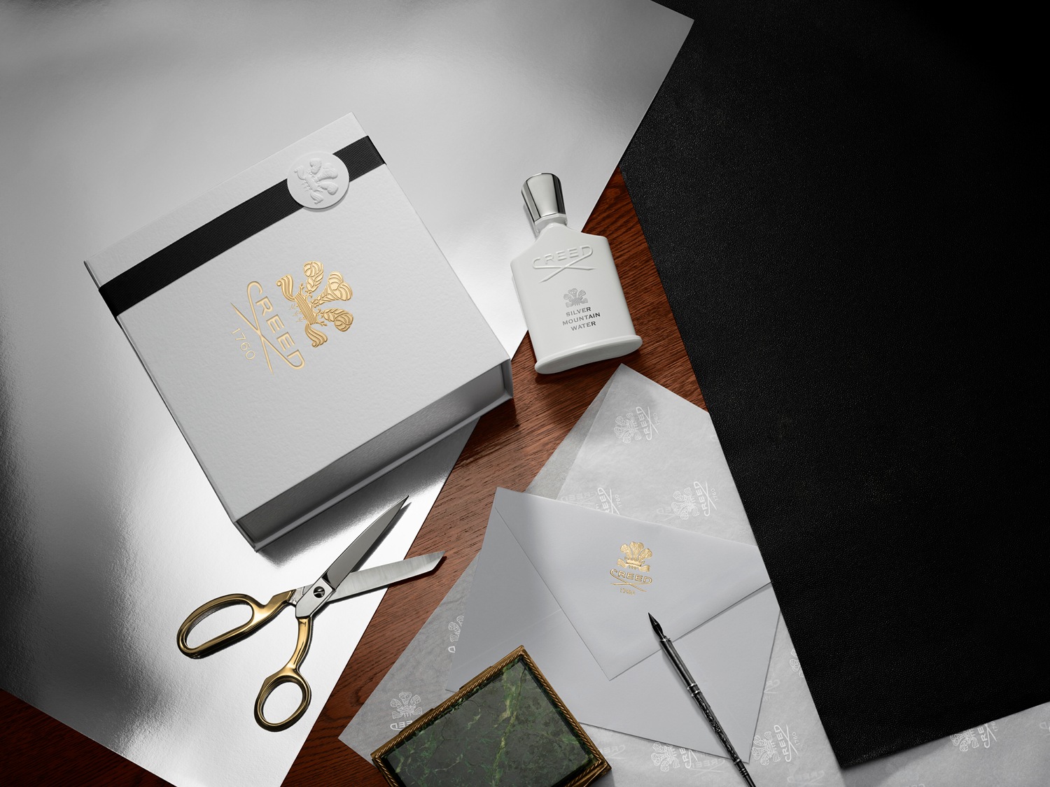 Elegantní sada Creed Silver Mountain Water s bílou krabičkou zdobenou zlatým logem, flakonem parfému, dopisním papírem a zlatými nůžkami vytváří luxusní atmosféru.