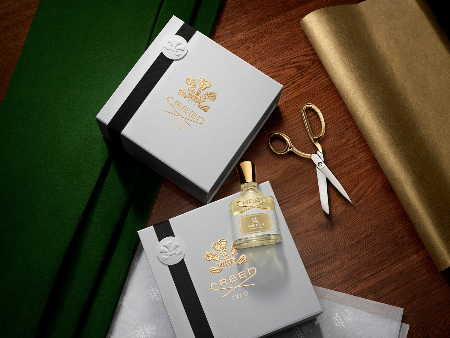A Creed Aventus for Her parfüm egy elegáns, szürke dobozban érkezik, arany logóval és fekete szalaggal, mellette arany olló és fényes papír látható.