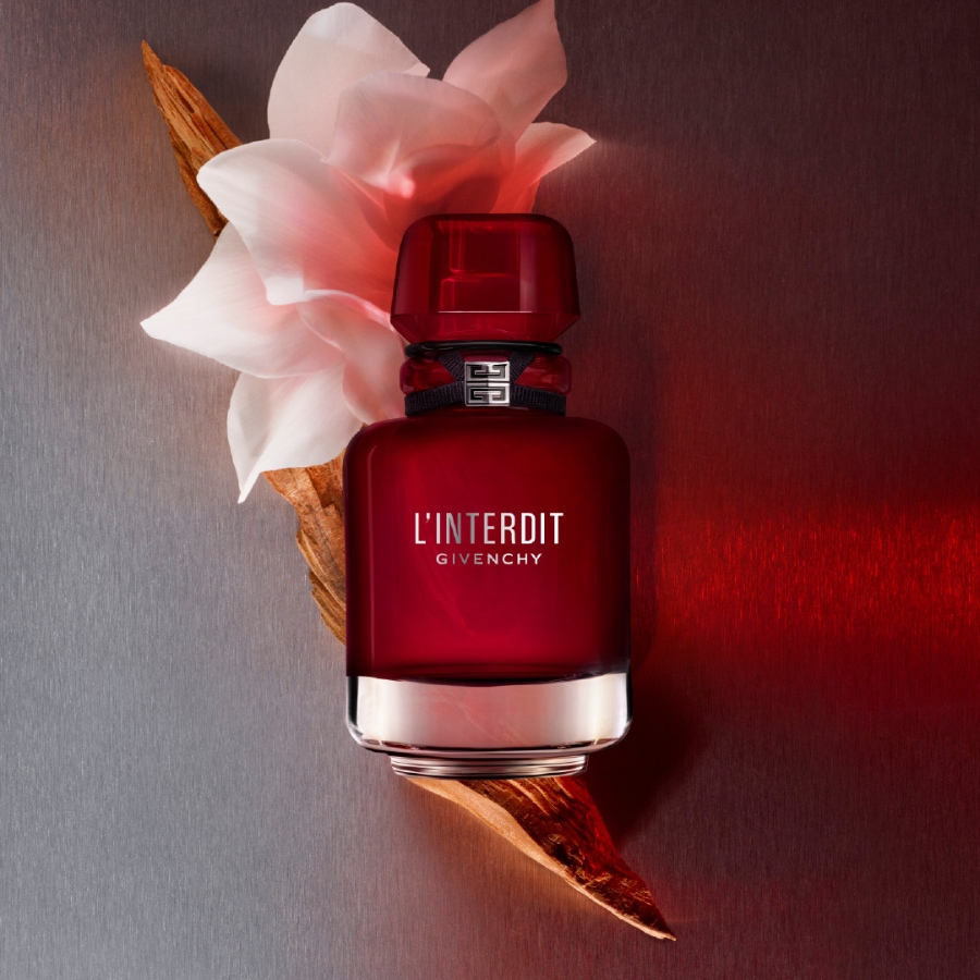 Der L'Interdit Givenchy Duft in einem eleganten, tiefroten Flakon mit silbernem Boden und schwarzem Band, verziert mit dem Givenchy-Logo, ruht auf einem Holzstück neben einer zarten weißen Blüte.