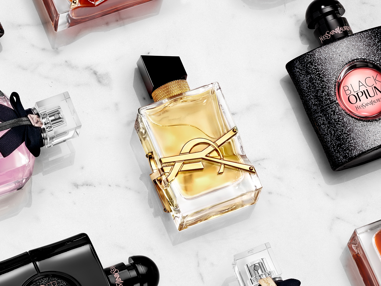 Na marmurowym tle leżą flakony perfum Yves Saint Laurent, w tym przezroczysty flakon z żółtą cieczą i złotymi elementami.