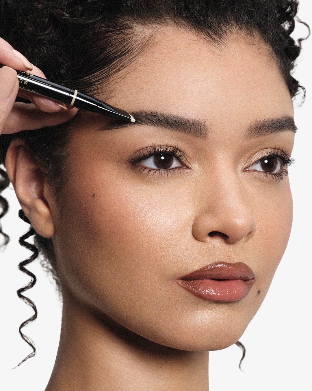 Nahaufnahme einer Frau mit dunklen Locken, die mit dem ArchiBrow Pencil von Anastasia Beverly Hills ihre Augenbrauen betont.