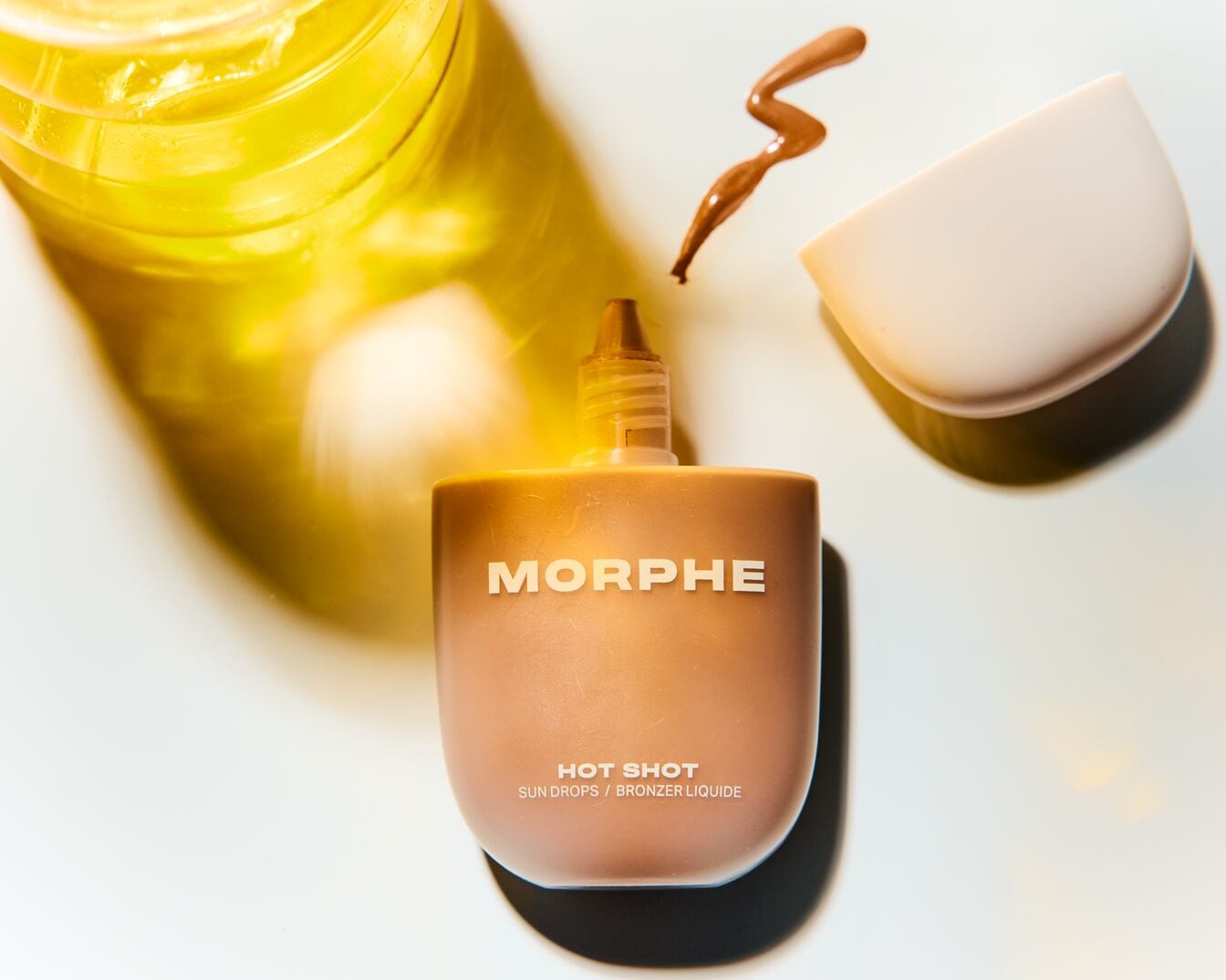 O sticlă bej de Morphe Hot Shot Sun Drops/Bronzer Liquide stă lângă o picătură de bronzer și un recipient deschis, oferind o strălucire însorită.