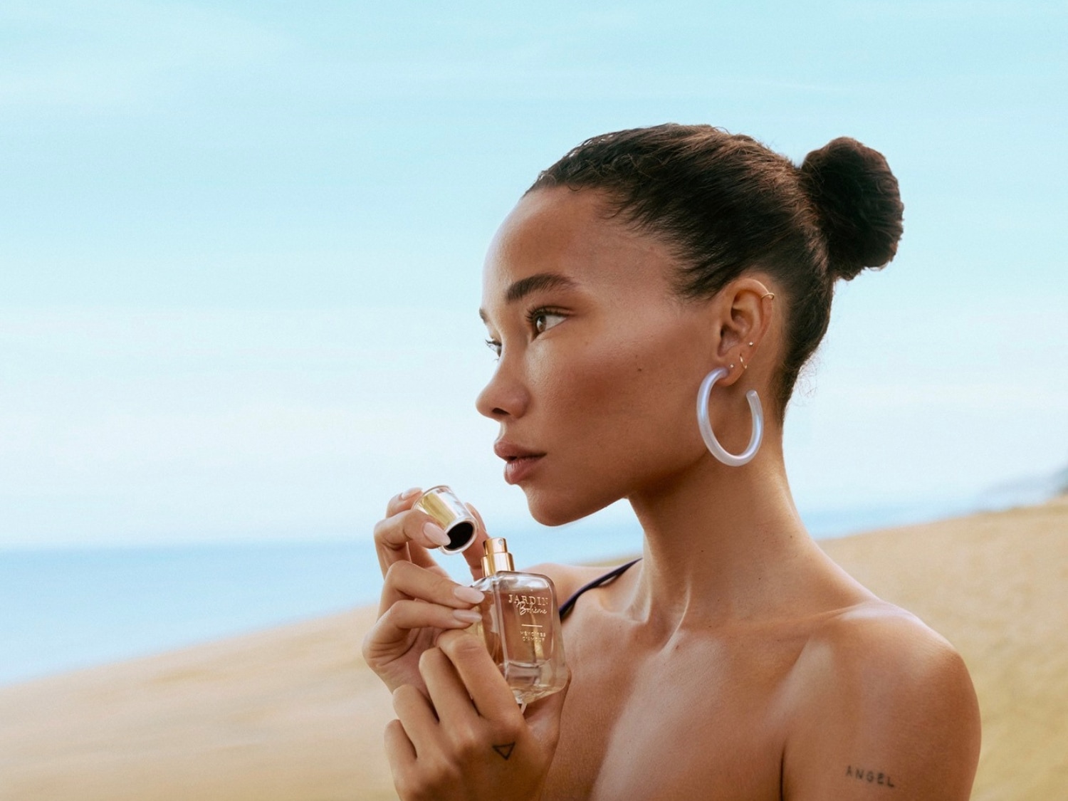Una donna con i capelli raccolti in uno chignon, indossa orecchini a cerchio bianchi e tiene in mano una bottiglia di profumo trasparente con tappo dorato, sullo sfondo una spiaggia e il cielo azzurro.