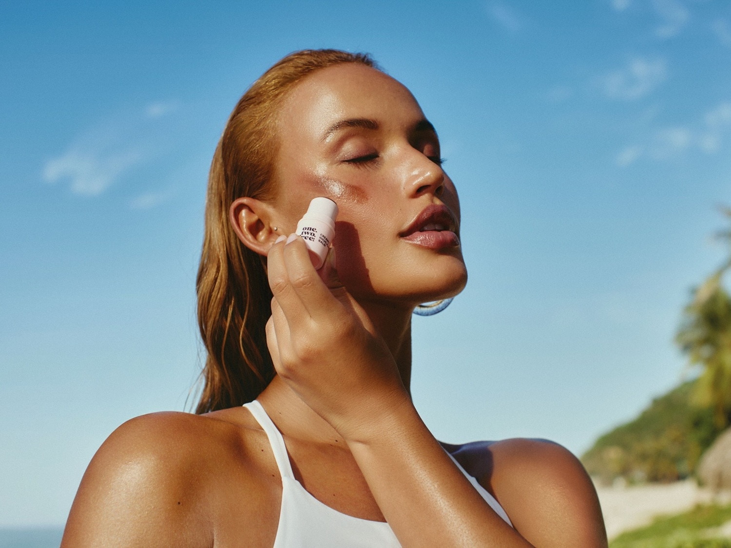 Una donna applica un fard in stick rosa sulla guancia, con il cielo azzurro e la spiaggia sullo sfondo, per un look fresco e luminoso.