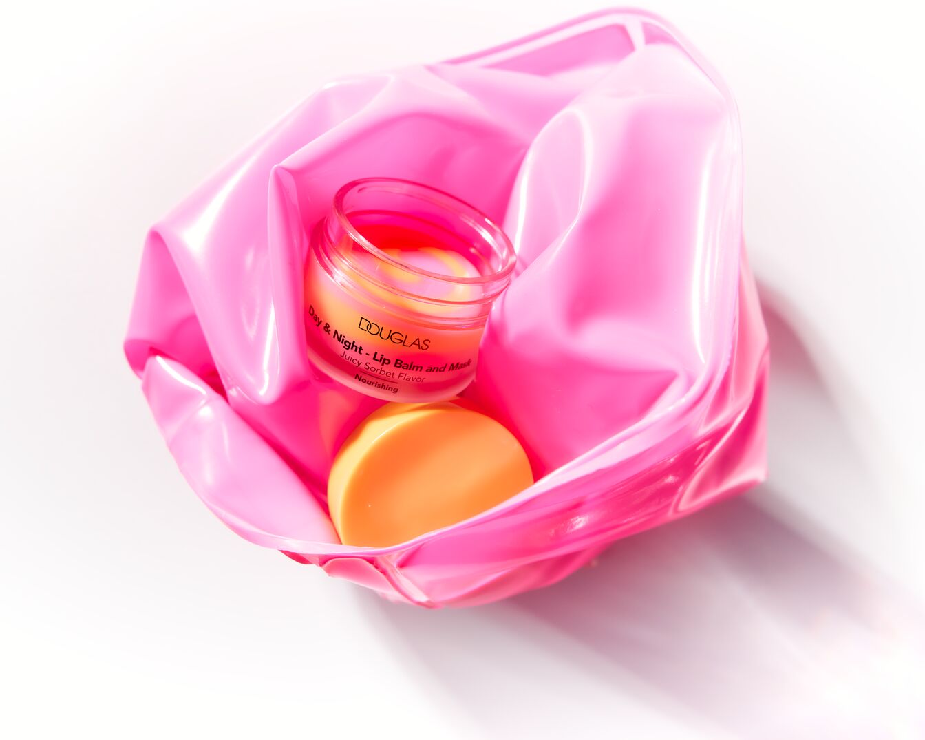 Balsamul de buze Douglas Day & Night într-un recipient transparent, cu o textură cremoasă în nuanțe de roz și galben, este prezentat într-o pungă roz lucioasă.