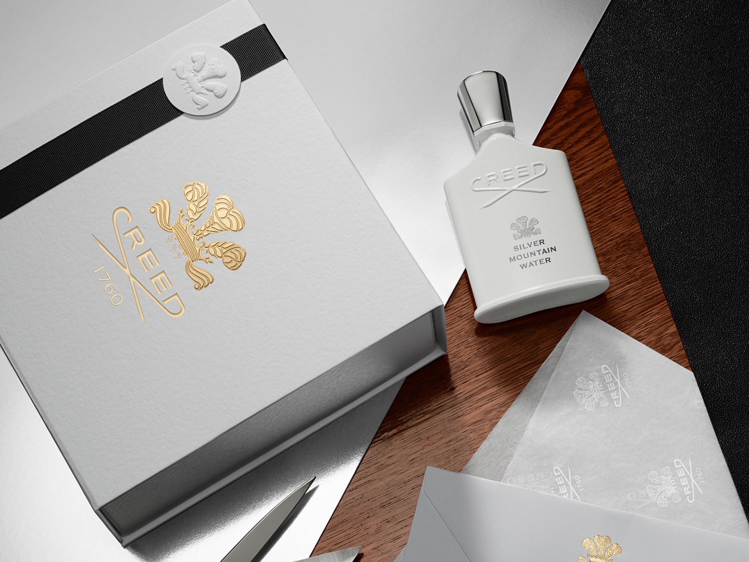 Elegancki zestaw upominkowy Creed Silver Mountain Water z białym pudełkiem ozdobionym złotym logo i czarną wstążką, obok flakon perfum na drewnianym stole.
