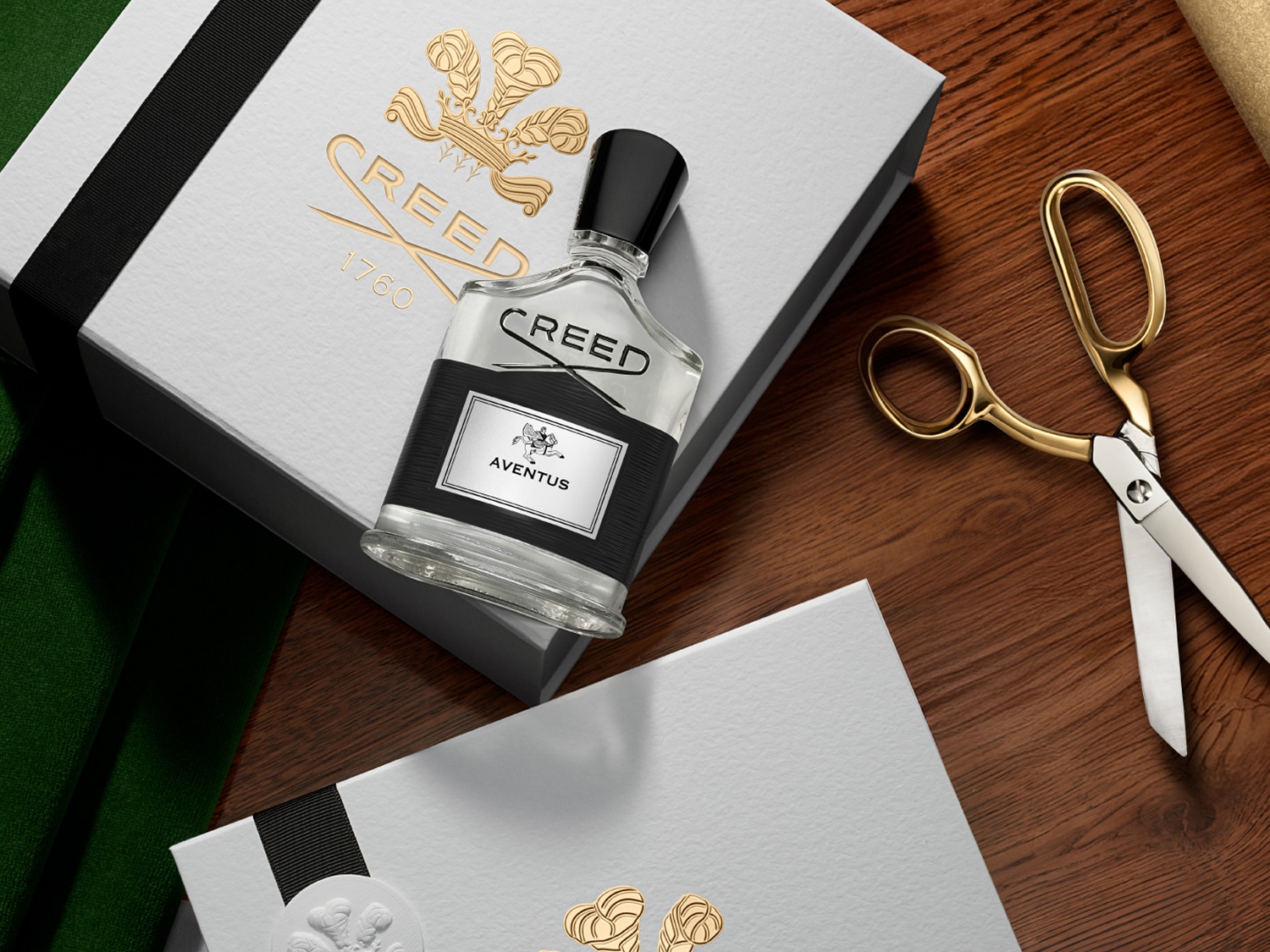 Elegancki flakon perfum Creed Aventus z czarną etykietą i korkiem, leżący na białym pudełku z logo marki w kolorze złota, obok złotych nożyczek na drewnianym stole.