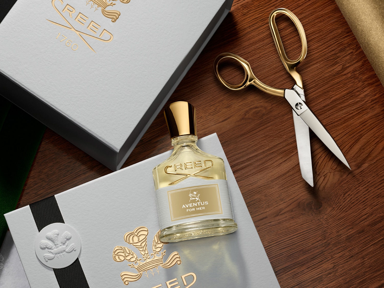 Butelka perfum Creed Aventus for Her stoi na pudełku obok złotych nożyczek na drewnianym stole, prezentując luksusowy zapach.